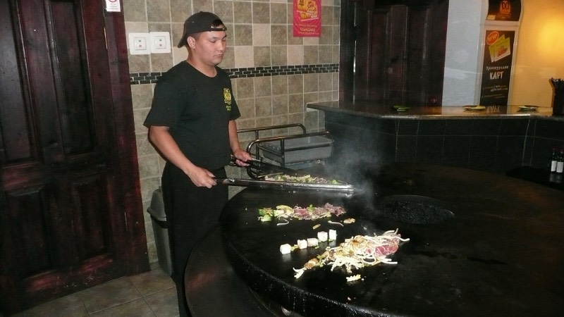 A cook stir-frying noodles and vegetables on a large round grill